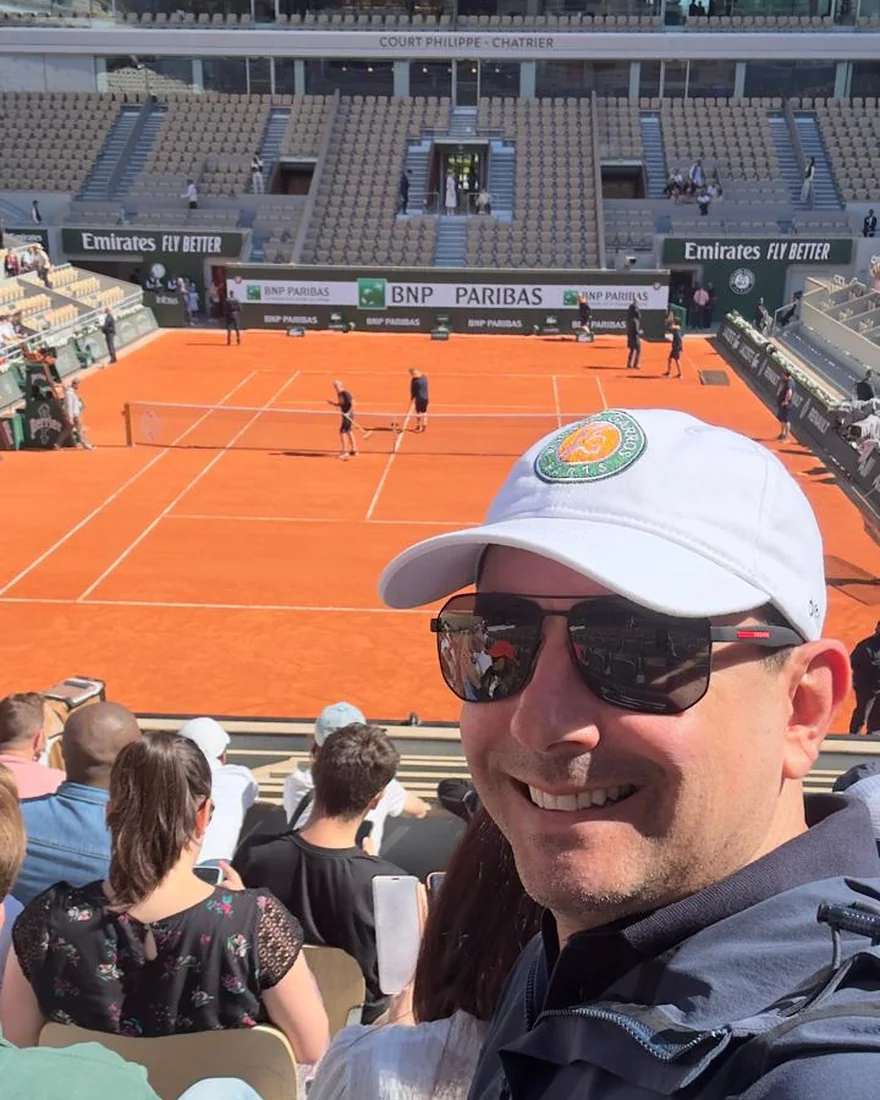 Diego Herling smiling in the stands of Court Philippe-Chatrier at Roland-Garros, Paris, in 2024, with the clay court behind him.